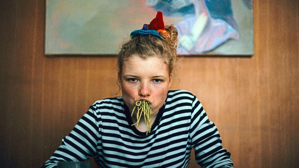 Einer jungen Frau in einem schwarz-weiß-geringelten T-Shirt und sommersprossigem Gesicht hängen Spaghetti mit grüner Sauce aus dem Mund.
