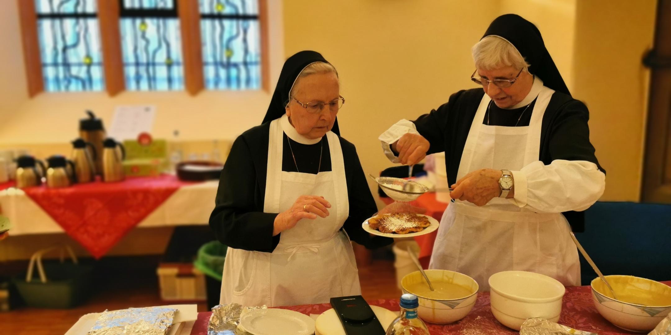 Zwei Ordensschwestern mit vorgebundener weißer Küchenschürze backen Herz-Waffeln und bestäuben diese mit Puderzucker.