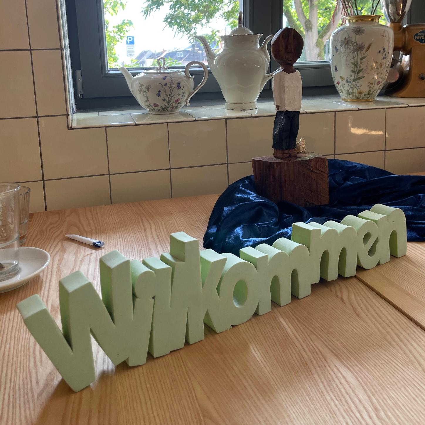 Die kleine Königsfigur, mit einem Arm, steht auf einem blauglänzendem Tuch hinter dem hellgrünen und dreidimensionalen Schriftzug 'Willkommen'.