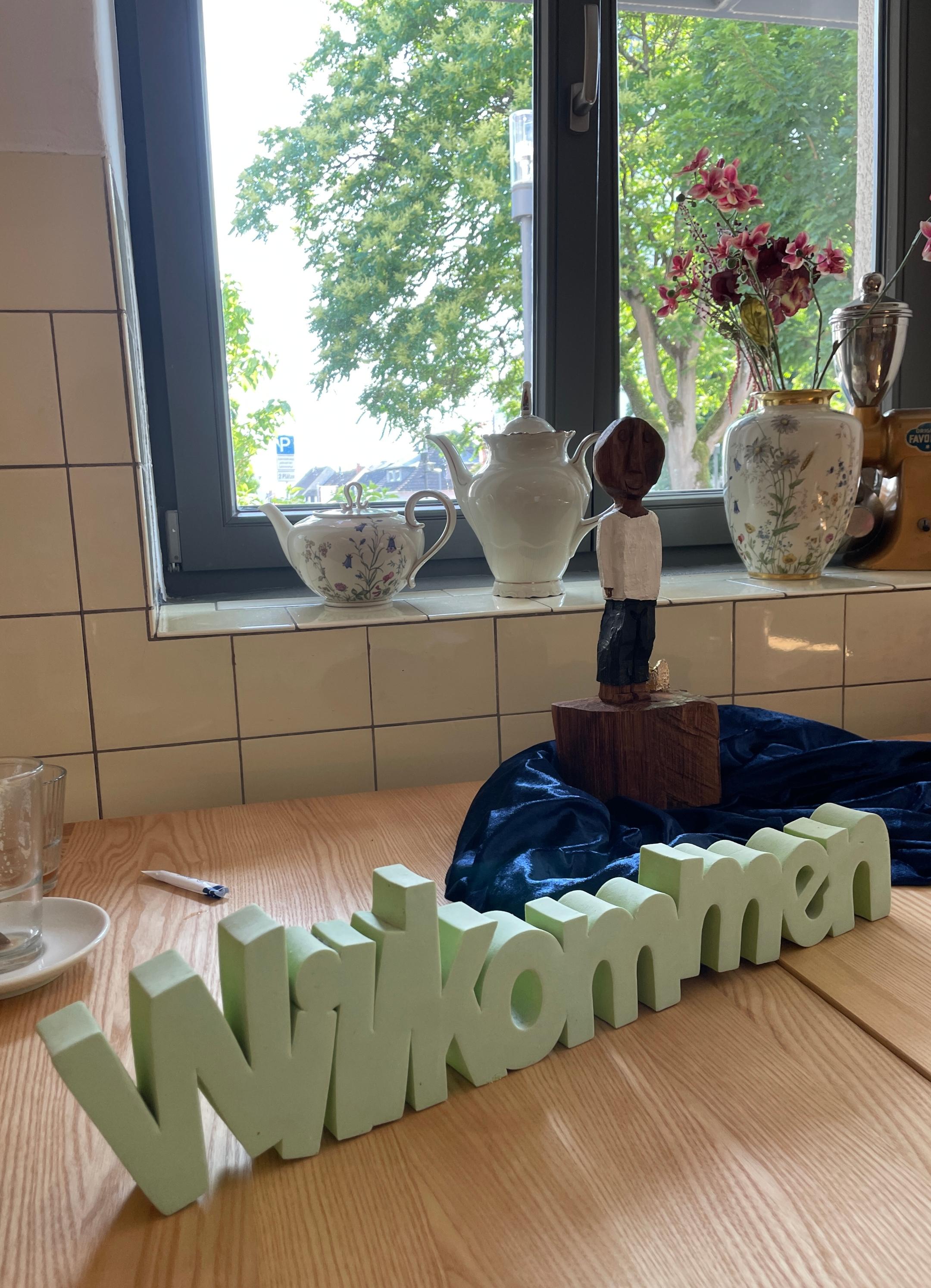 Die kleine Königsfigur, mit einem Arm, steht auf einem blauglänzendem Tuch hinter dem hellgrünen und dreidimensionalen Schriftzug 'Willkommen'.