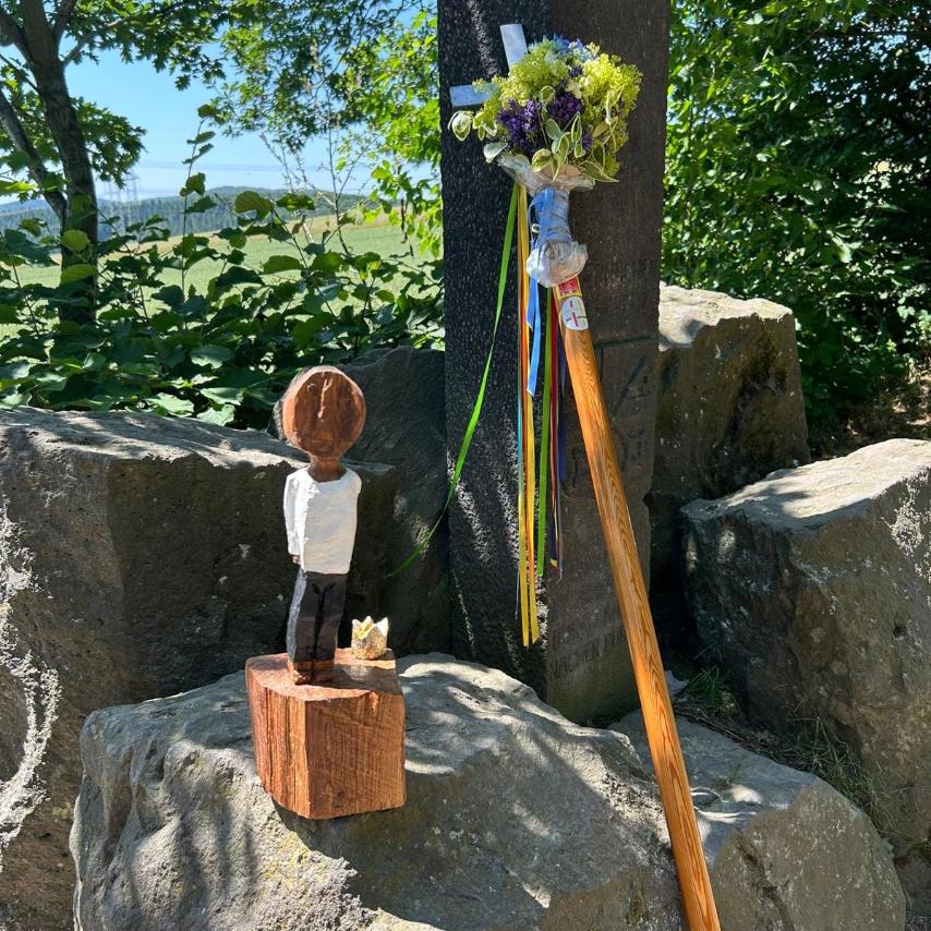 Die kleiner Königsfigur steht auf einem Felsblock in freier Natur. Daneben lehnt das mit Blumen geschmückte Pilgerkreuz an einem Baum.