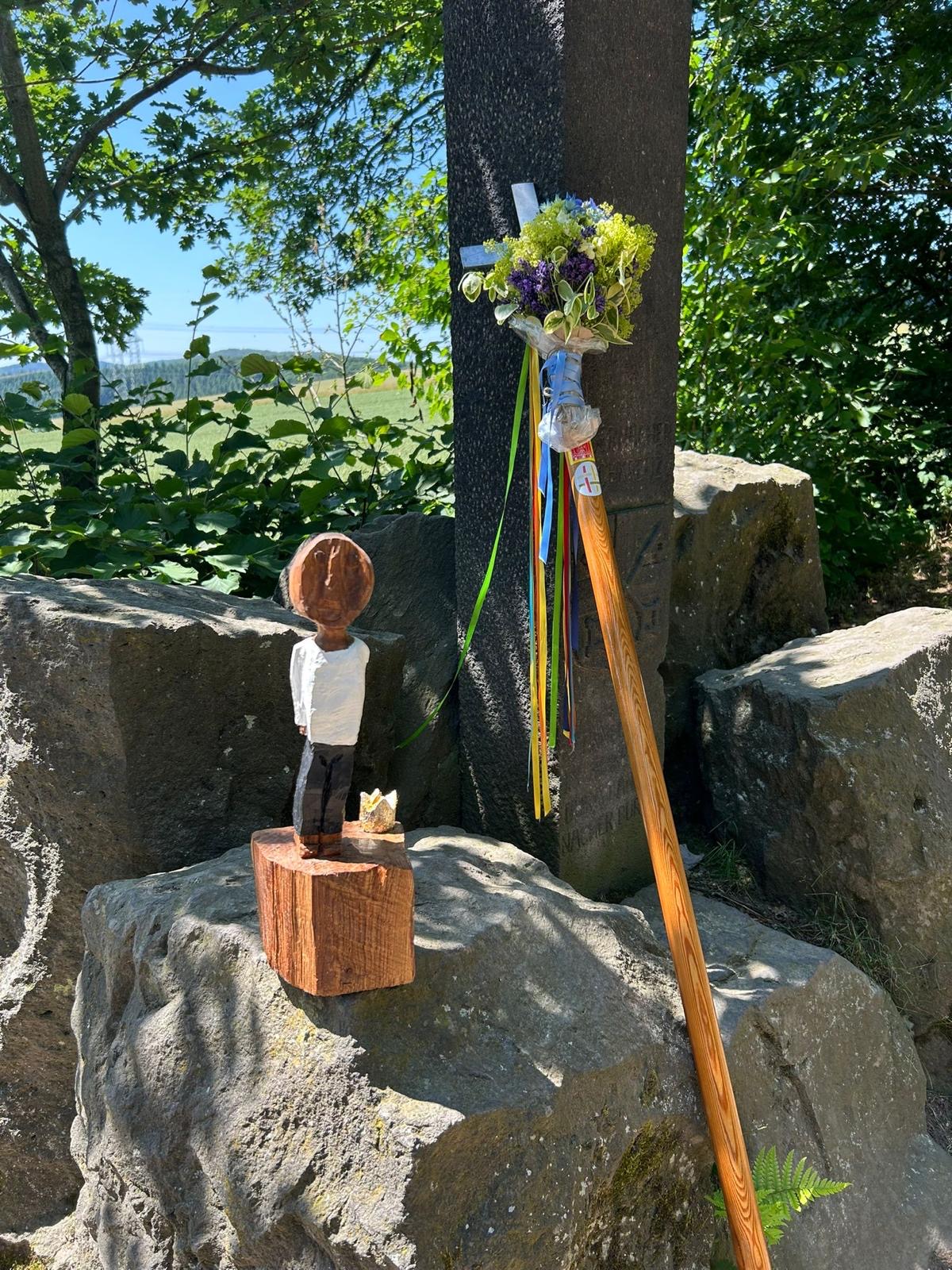 Die kleiner Königsfigur steht auf einem Felsblock in freier Natur. Daneben lehnt das mit Blumen geschmückte Pilgerkreuz an einem Baum.