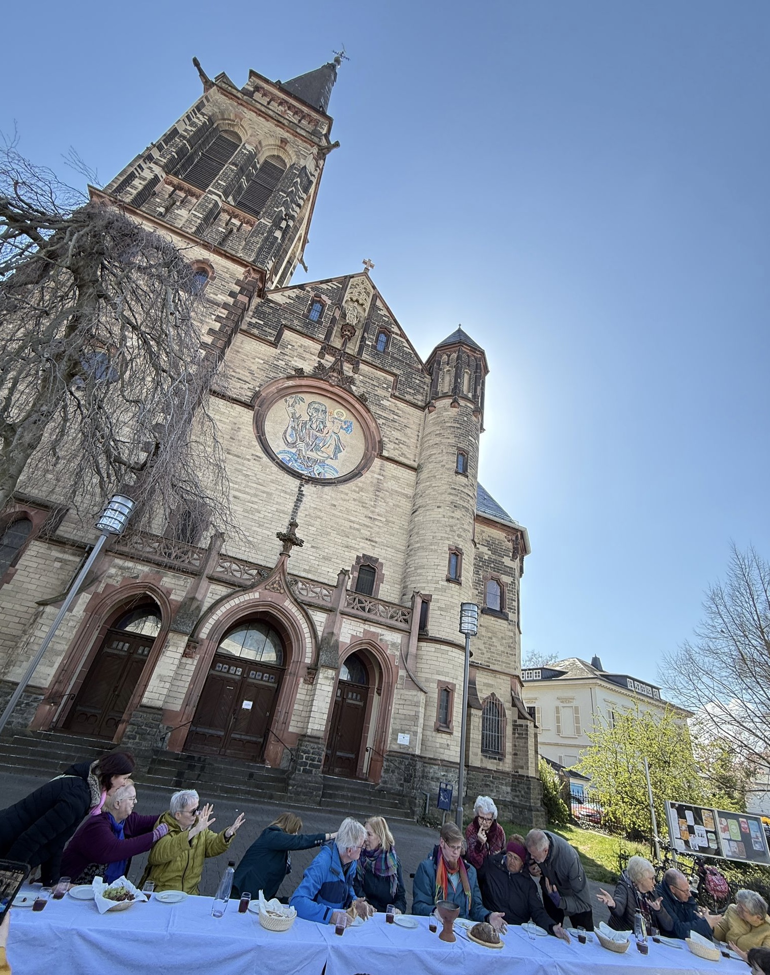 Die 13 Laienschauspieler*innen sitzen vor der Matthias-Kirche an einer langen, mit weißen Tischdecken gedeckten Tafel.