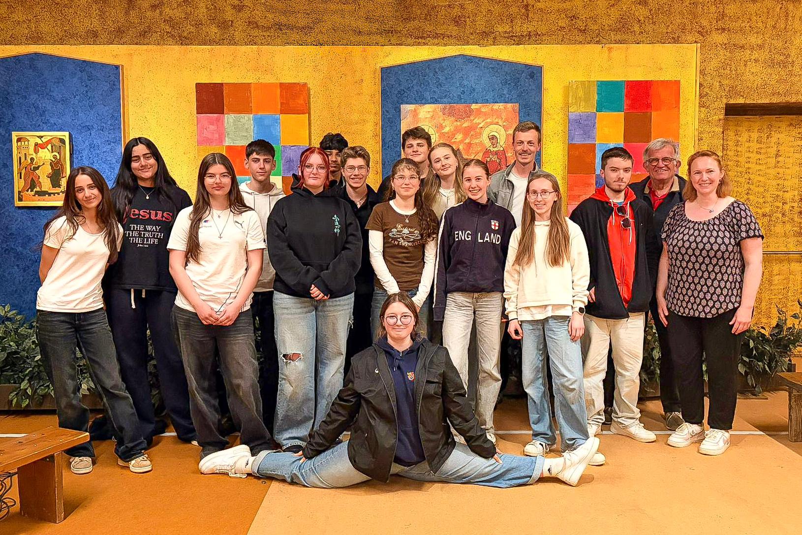 Die Reisegruppe, 17 Personen, vor allem Jugendliche steht im Altarraum in Taizé vor einem bunten Hintergrund, u. a. mit zwei Ikonen, und lächelt in die Kamera. Eine Teilnehmerin sitzt im Damenspagat vor der Gruppe.