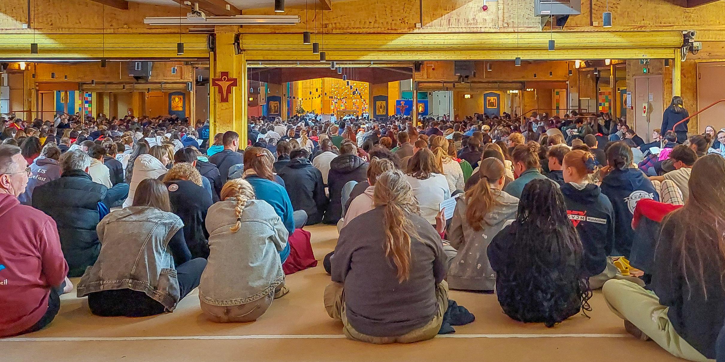 Der Blick geht über die Rücken der auf den Boden sitzenden meist jungen Menschen hin zum Chorraum der Kirche. An den Wänden hängen vereinzelt Ikonen vor blauem Hintergrund.