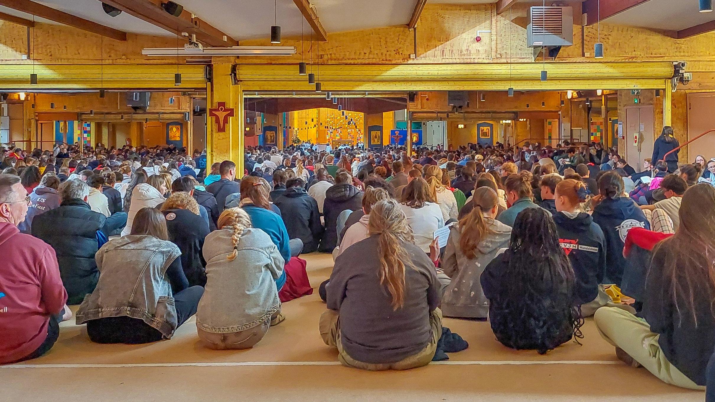 Der Blick geht über die Rücken der auf den Boden sitzenden meist jungen Menschen hin zum Chorraum der Kirche. An den Wänden hängen vereinzelt Ikonen vor blauem Hintergrund.
