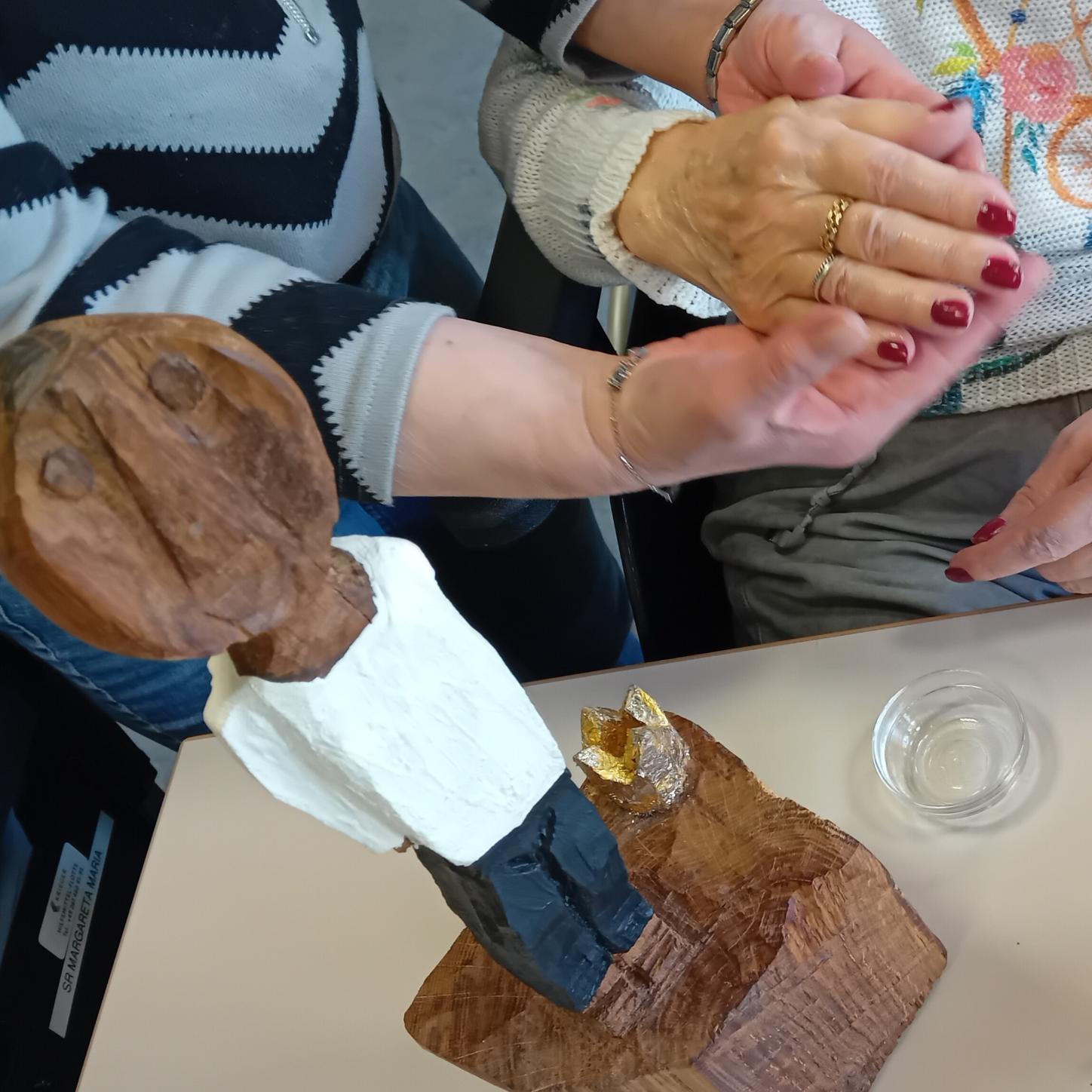 Die kleine Königsfigur steht auf einem Tisch. Daneben werden alte Hände von jüngeren gesalbt.