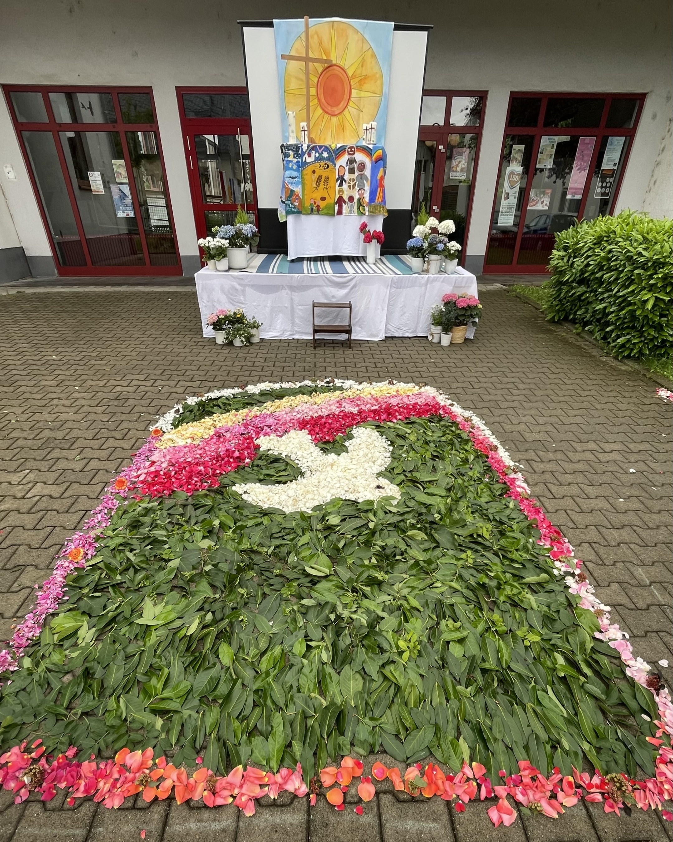 Hinter den mit Blüten und Blättern gestalteten Heilig-Geist-Bild, das einen kleinen Regenbogen und eine weiße Taube zeigt, ist der Altar zu sehen, den Kommunionkinder mit Hortensien, selbstgemalten Bildern, selbstgestalteten Kerzen und einem großen Bild einer Sonne gestaltet haben.