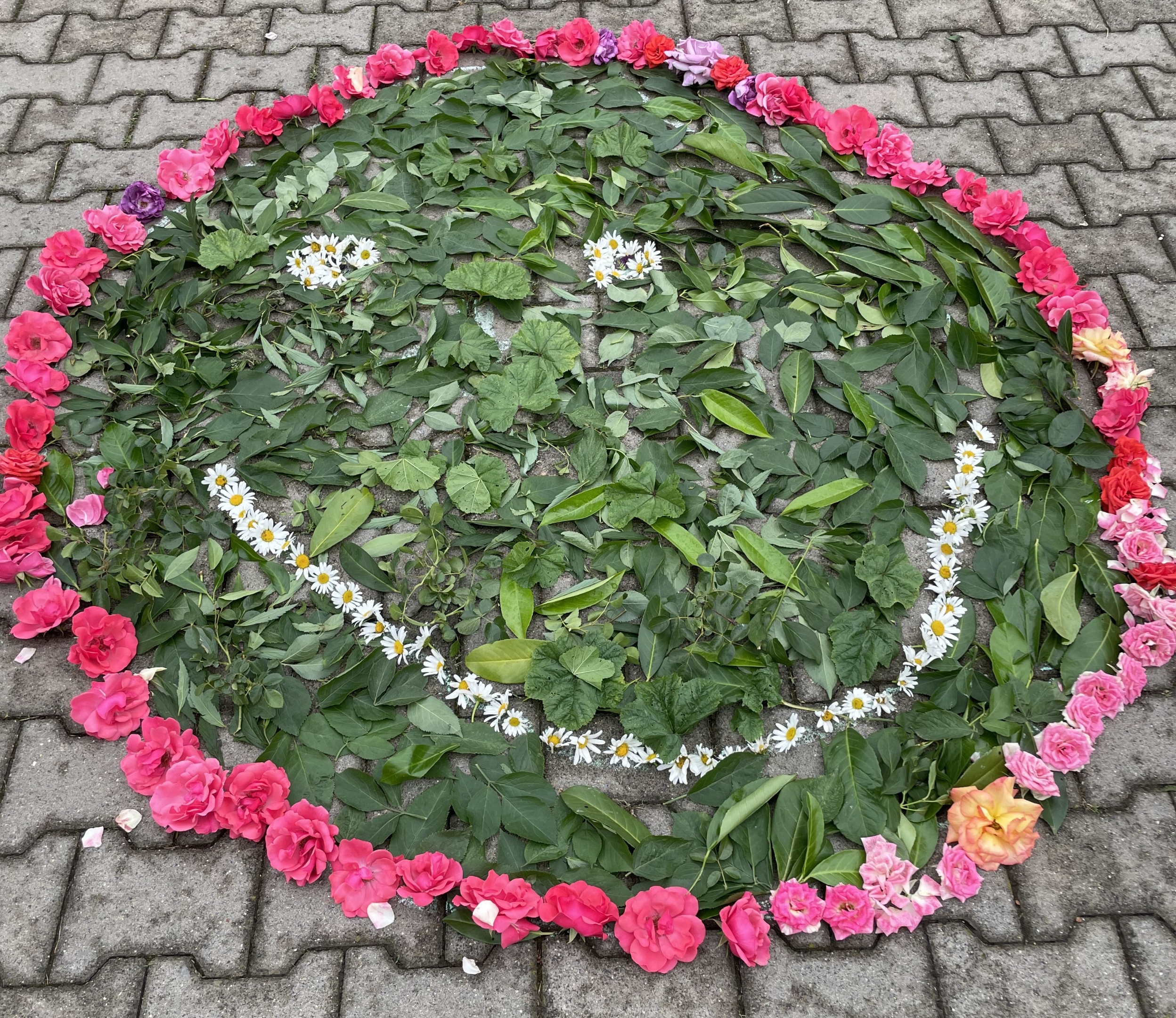 Rote Rosenblüten bilden auf grauen Pflastersteinen den Rand des Smileys, der mit grünen Blättern gefüllt ist. Die Augen und der lachende Mund sind durch Gänseblümchen dargestellt.