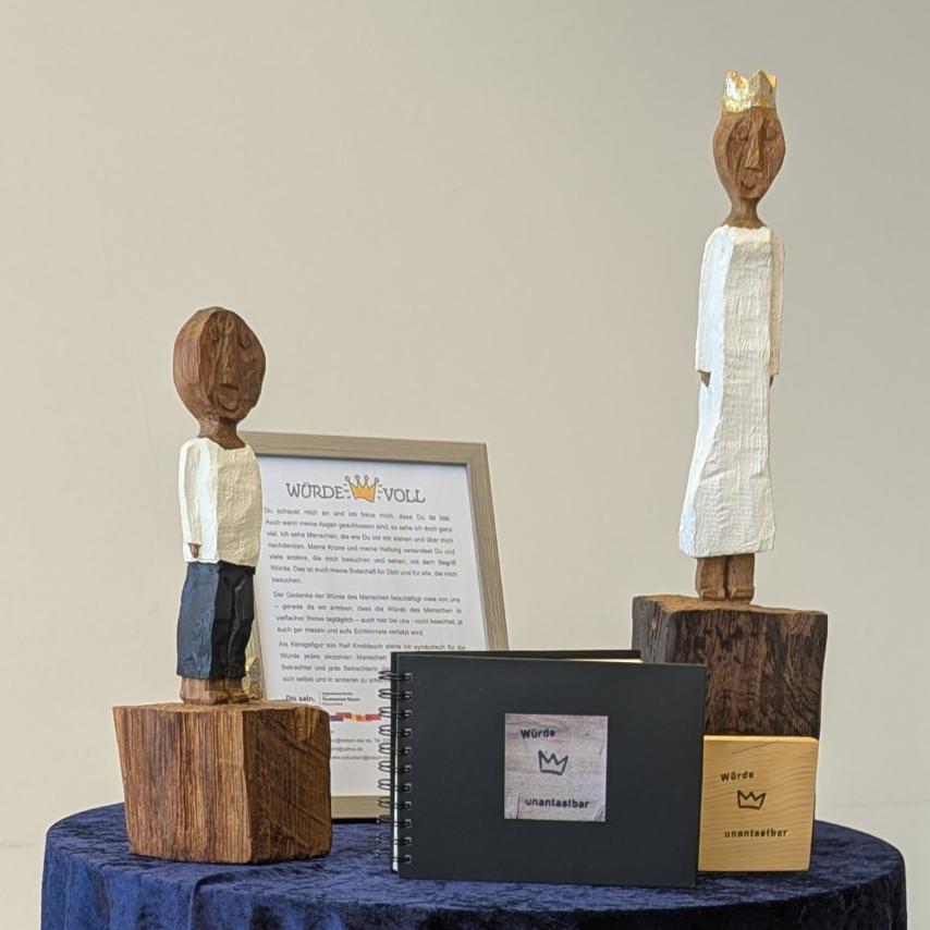 Die beiden Königsfiguren stehen auf einem mit einem blauen Samttuch gedeckten Stehtisch, zusammen mit einem erläuternden Aufsteller, einer  'Würde-unantastbar'-Tafel und ihrem Reisebuch.