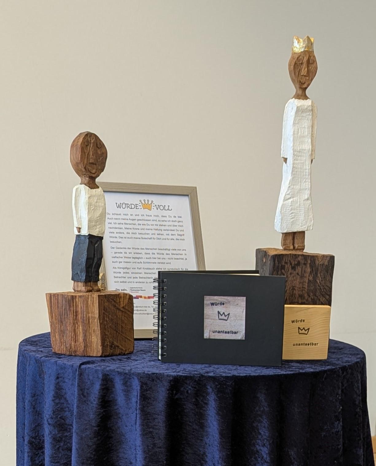 Die beiden Königsfiguren stehen auf einem mit einem blauen Samttuch gedeckten Stehtisch, zusammen mit einem erläuternden Aufsteller, einer  'Würde-unantastbar'-Tafel und ihrem Reisebuch.