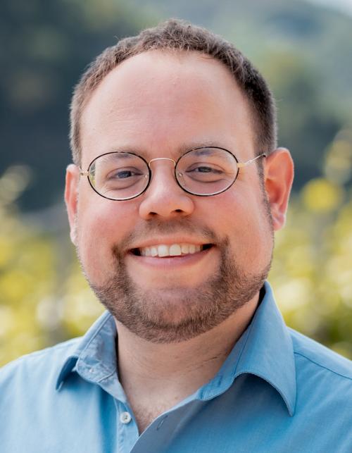 Ein Mann, Ende 30, schaut lächelnd in die Kamera. Eine hohe Stirn schließt sich an seine Nickel-Brille an. Er trägt einen dunkelbraunen gestutzten Vollbart.