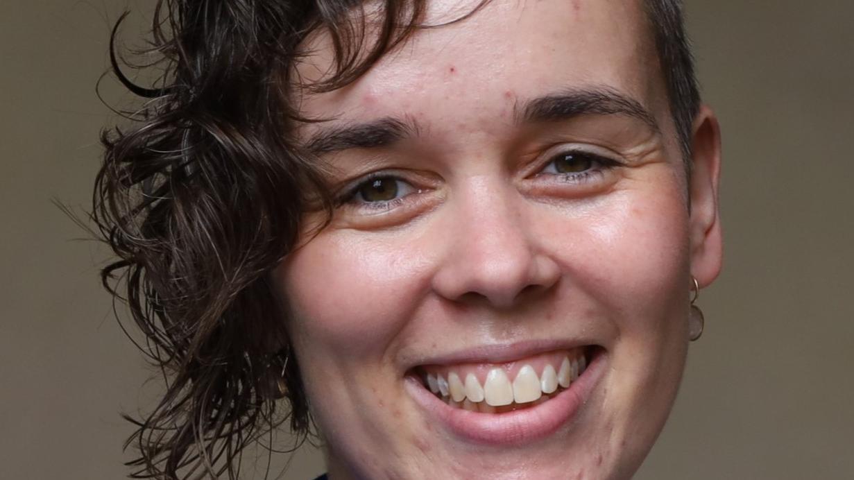 Eine junge Frau mit einem ovalen Gesicht und dunkelbraunen Haaren, auf ihrer rechten Seite lang, auf der anderen kurzgeschnitten, strahlt Sie an.
