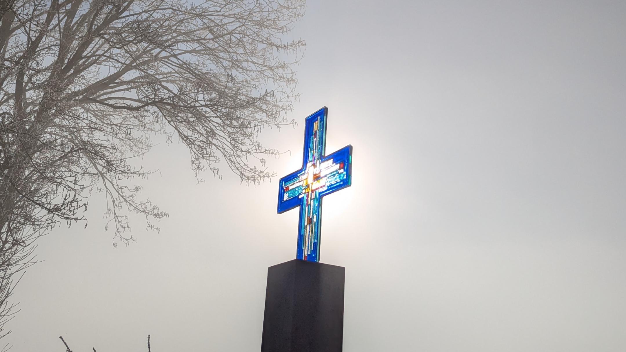 Ein blaues Glaskreuz, mit wenigen anderen Farben, steht auf einer Steinstele vor einem Feld und wird von der vernebelten Wintersonne von hinten beleuchtet.