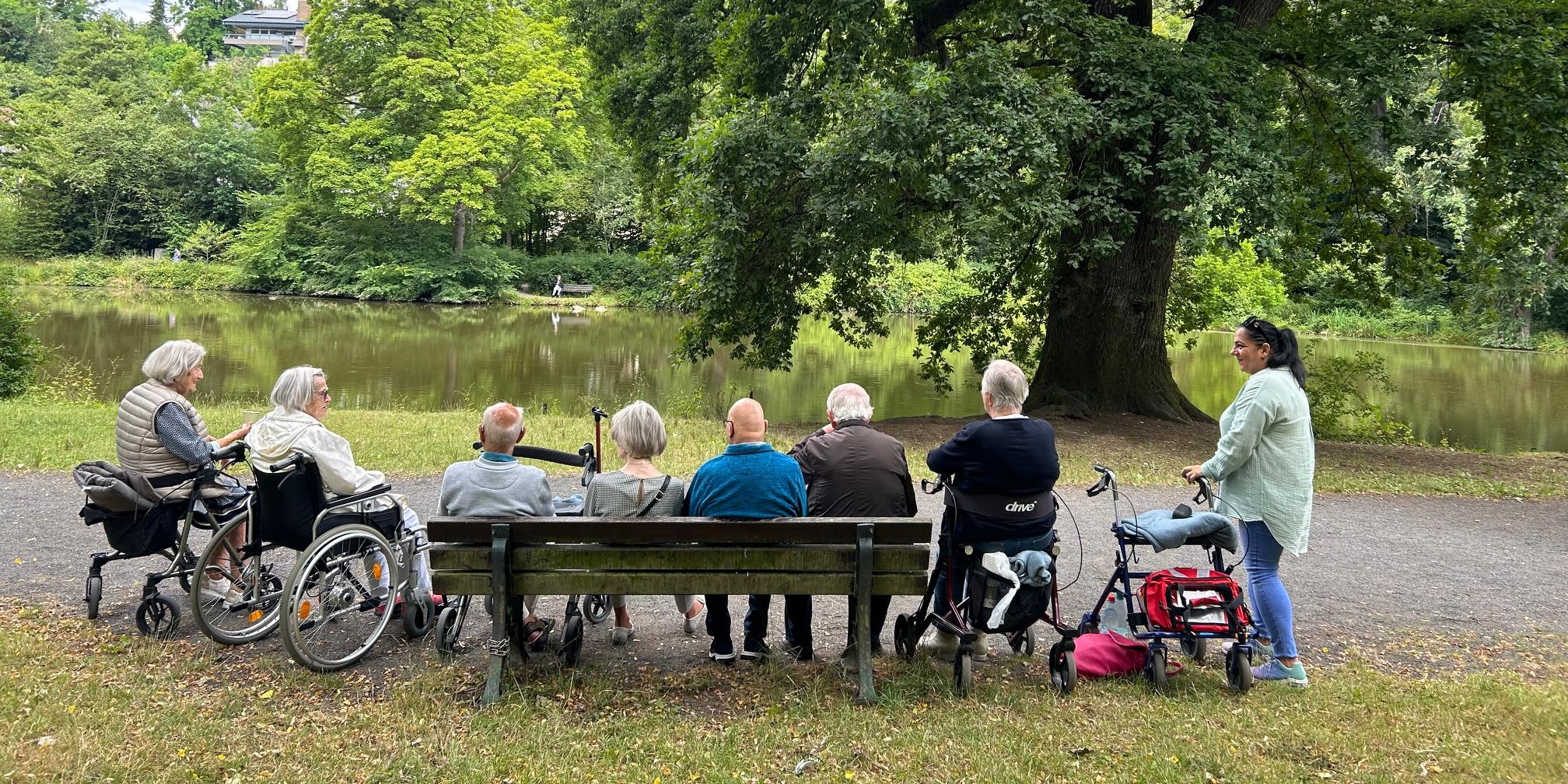 Sieben Senioren*innen sitzen auf einer Bank, im Rollstuhl, auf Rollatoren am Teich im Schlosspark Sayn und schauen aufs Wasser.