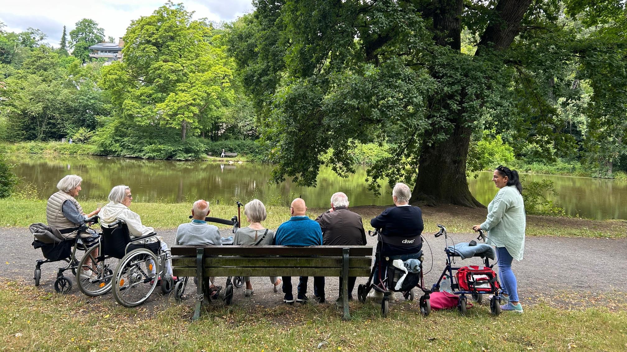 Sieben Senioren*innen sitzen auf einer Bank, im Rollstuhl, auf Rollatoren am Teich im Schlosspark Sayn und schauen aufs Wasser.