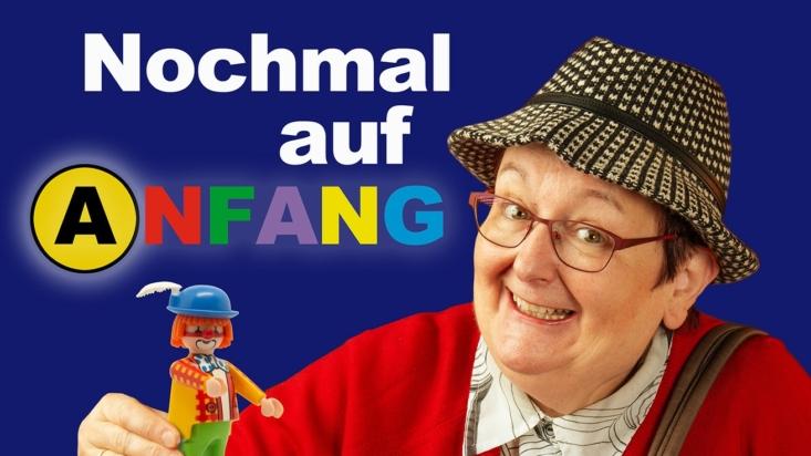 Eine ältere Dame in roter Jacke und Pepita-Hut sitzt hinter einem Mensch-Ärgere-Dich-Nicht-Brett, das mit unterschiedlichen Playmobilfiguren bestellt ist.