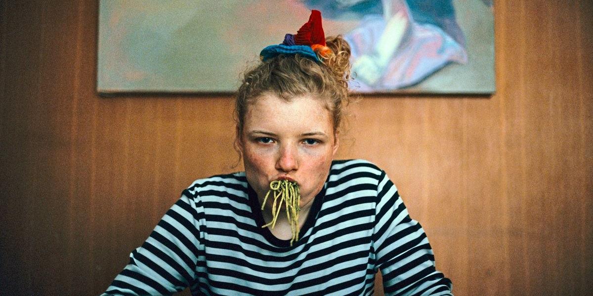 Einer jungen Frau in einem schwarz-weiß-geringelten T-Shirt und sommersprossigem Gesicht hängen Spaghetti mit grüner Sauce aus dem Mund.