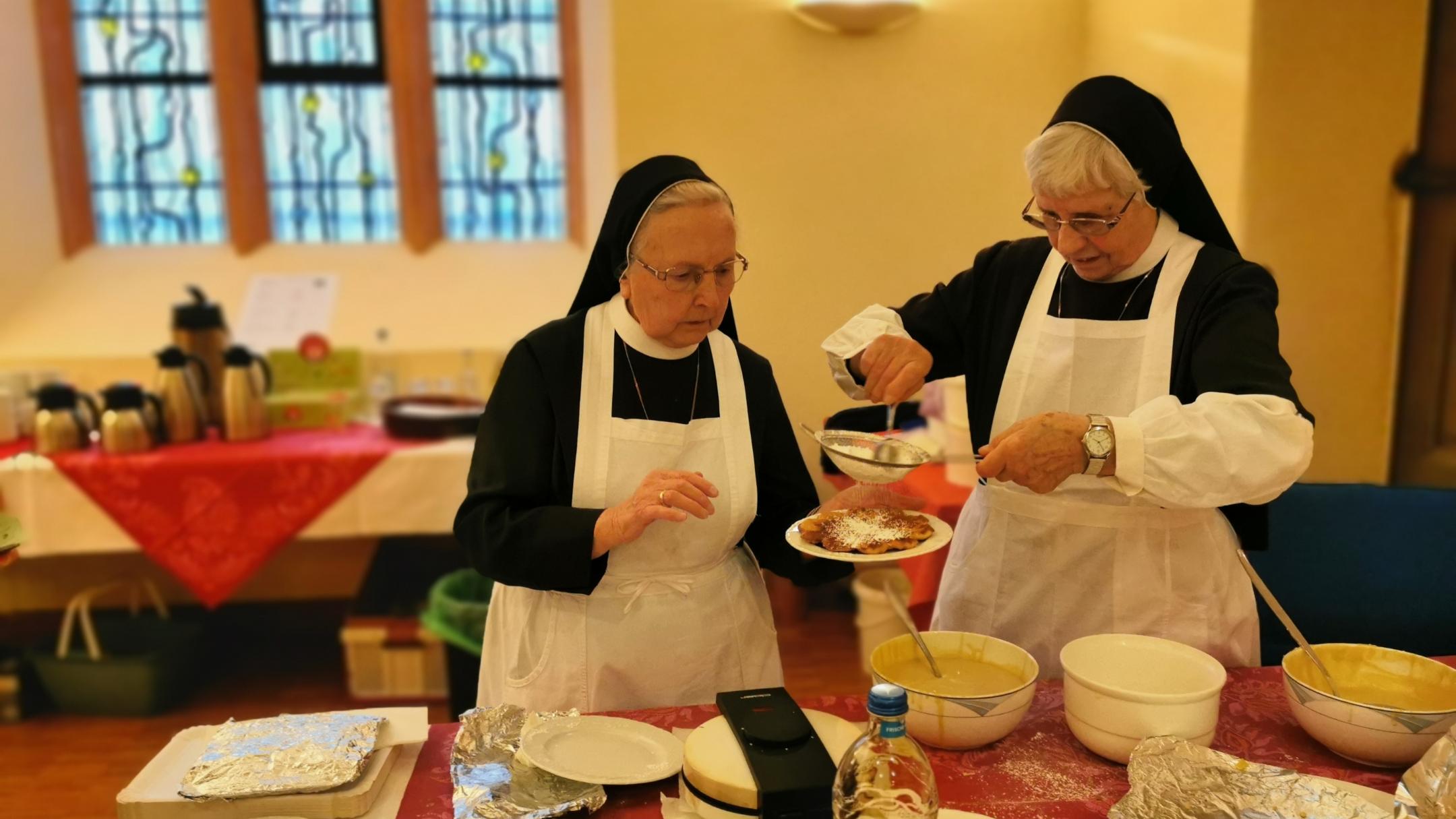 Zwei Ordensschwestern mit vorgebundener weißer Küchenschürze backen Herz-Waffeln und bestäuben diese mit Puderzucker.