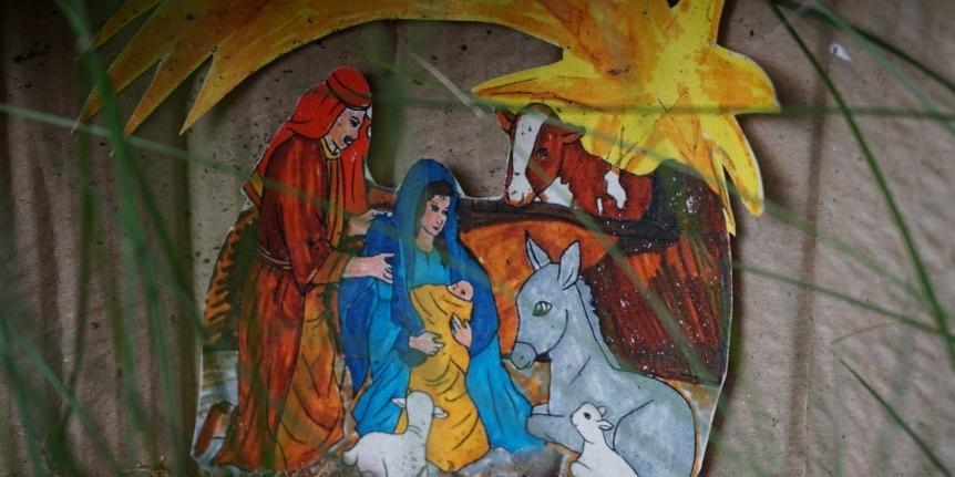 Eine colorierte Krippendarstellung mit Josef, Maria, Jesus-Baby, Ochs, Esel, zwei Schafen und dem Weihnachtsstern ist hinter Grashalmen vor Betonsteinen zu sehen.