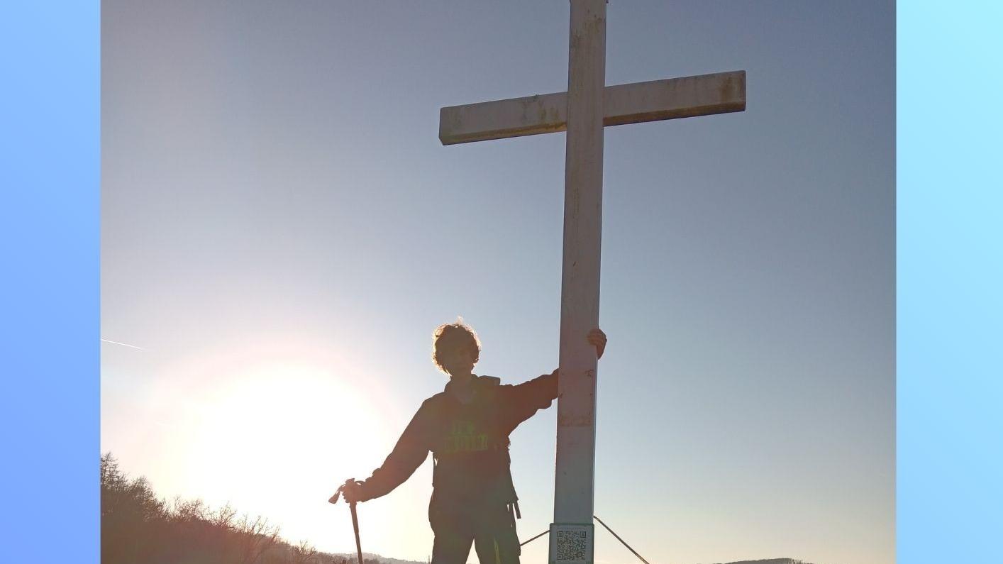 Im Gegenlicht steht ein junger Mann auf einem Berg an einem Gipfelkreuz.