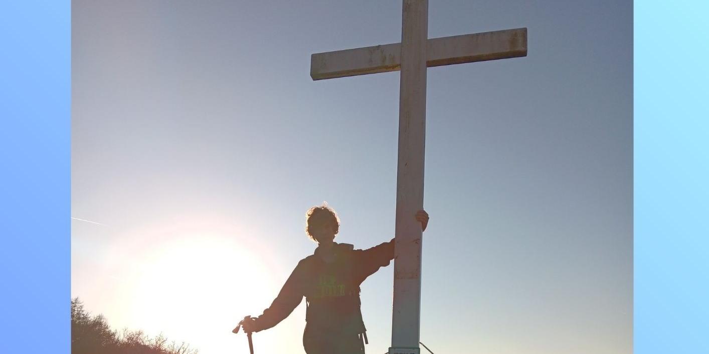 Im Gegenlicht steht ein junger Mann auf einem Berg an einem Gipfelkreuz.