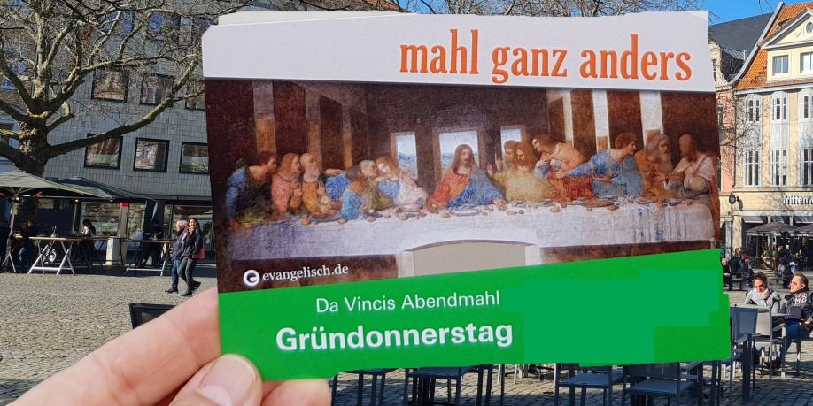 Eine Hand hält eine Postkarte mit dem Motiv des letzten Abendmahls von da Vinci, im Hintergrund ein Marktplatz.