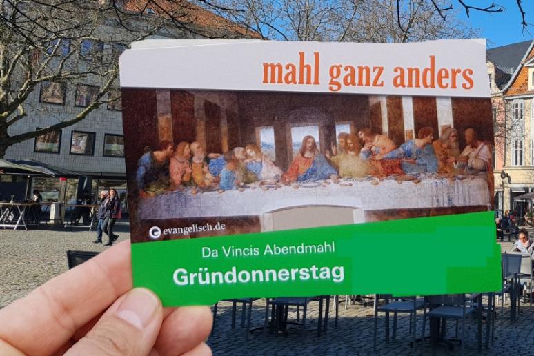 Eine Hand hält eine Postkarte mit dem Motiv des letzten Abendmahls von da Vinci, im Hintergrund ein Marktplatz.