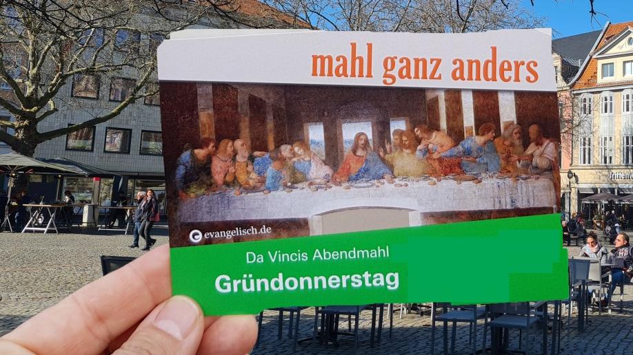 Eine Hand hält eine Postkarte mit dem Motiv des letzten Abendmahls von da Vinci, im Hintergrund ein Marktplatz.