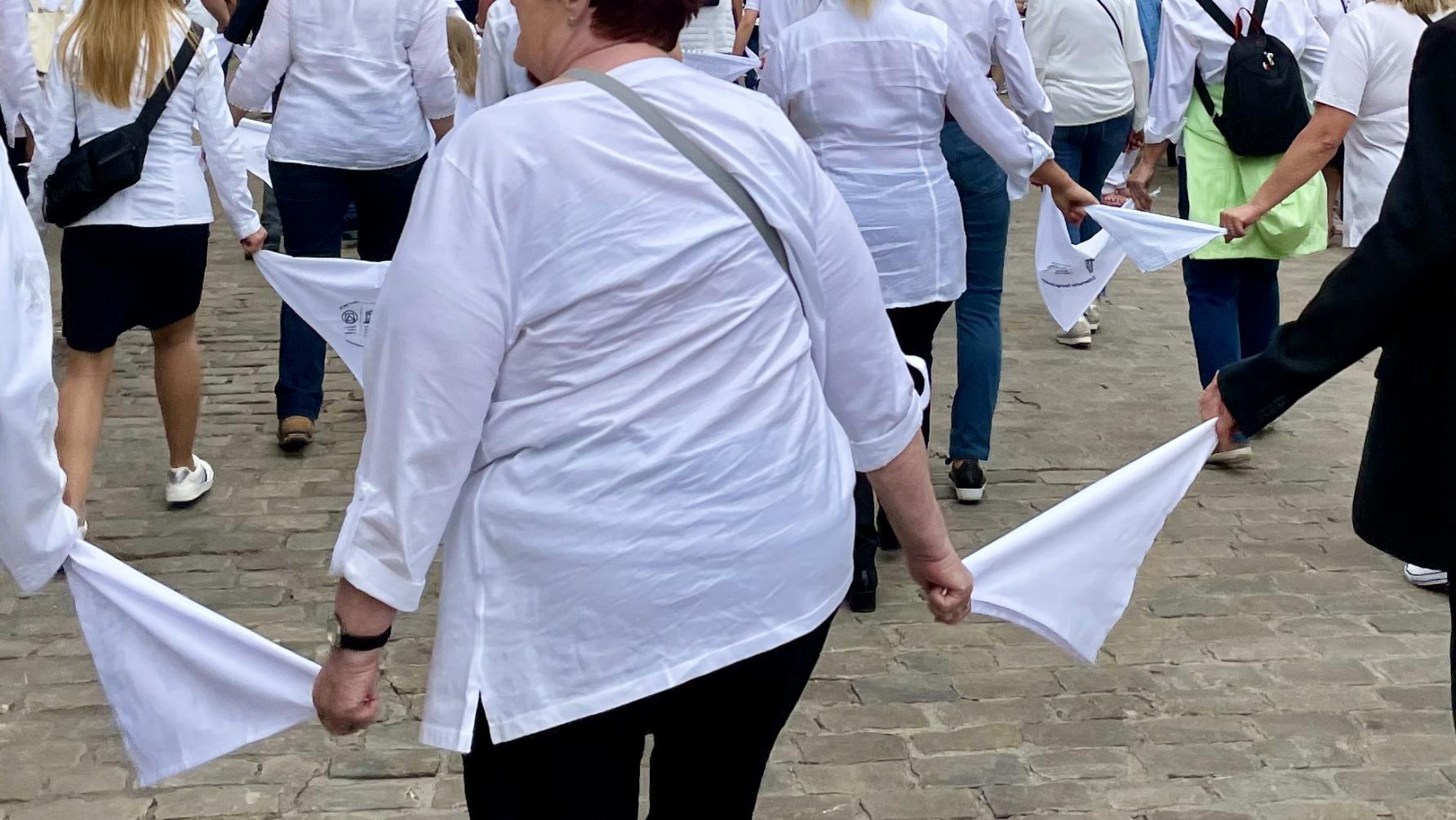 Das Bild zeigt Menschen in dunklen Hosen und weißen Oberteilen, die jeweils durch ein weißes Tuch verbunden in Reihen durch die Straßen 'springen'.