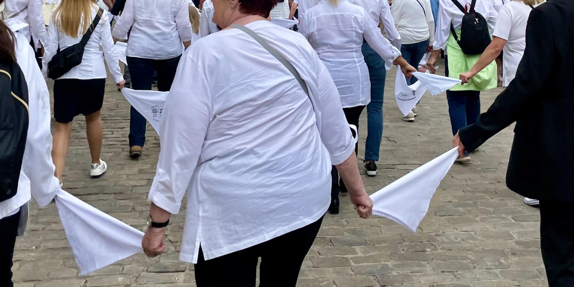 Das Bild zeigt Menschen in dunklen Hosen und weißen Oberteilen, die jeweils durch ein weißes Tuch verbunden in Reihen durch die Straßen 'springen'.