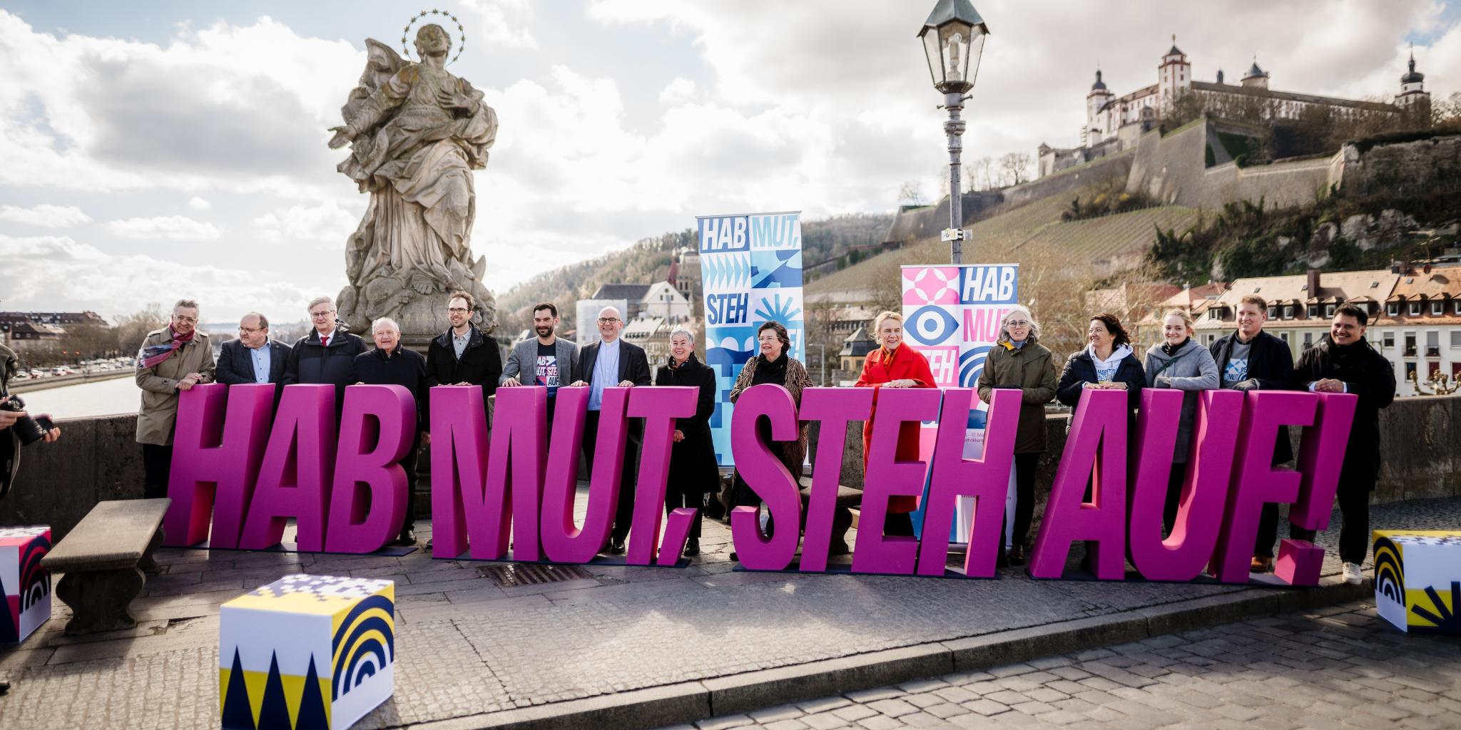 15 Menschen halten auf der Alten Mainbrücke die violetten Einzel-Großbuchstaben des Katholikentagsmottos: 'Hab Mut, steh auf!'