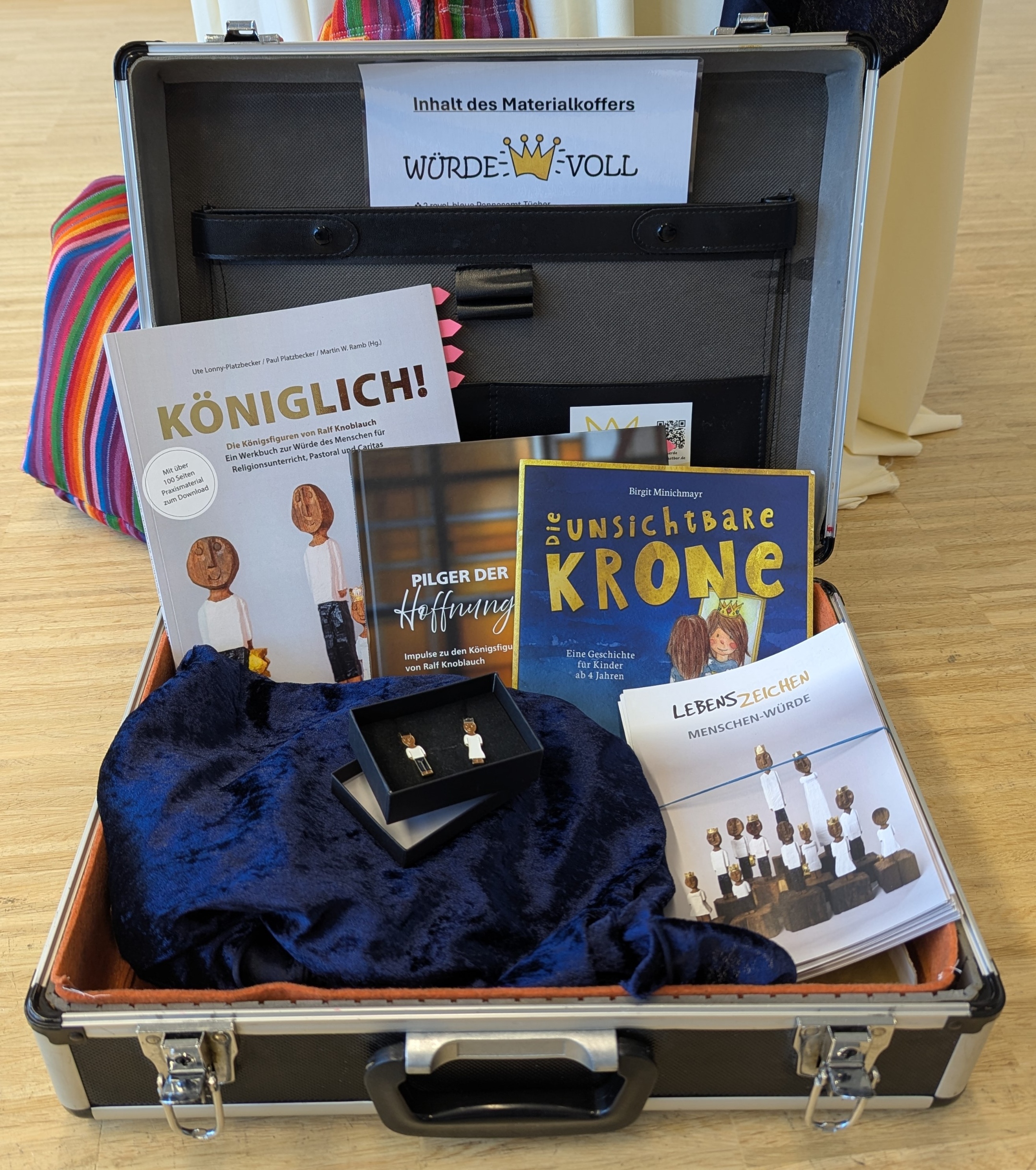 Der Materialkoffer aus Metall enthält zwei blaue Samttücher, Ansteckpins, Broschüren und Bücher.