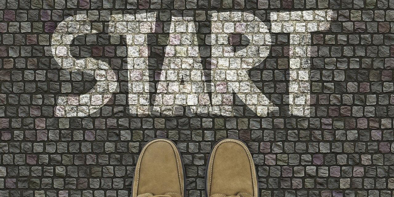 Auf kleinen Pflastersteinen ist mit Kreide das Wort 'Start' geschrieben, Fußspitzen stehen darunter.